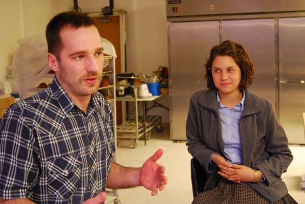 A young man and woman in a baking facility, sitting on stools, talking