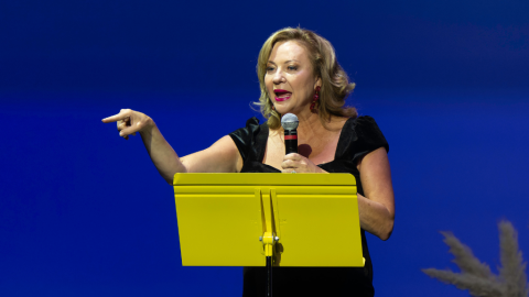Woman in black dress on stage behind a yellow stand point to crowd. 